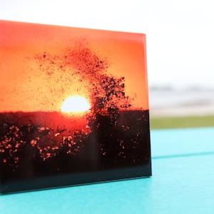 Pode incluir: Um azulejo de cerâmica com uma fotografia de um pôr do sol sobre uma massa de água. O sol está se pondo à distância, e a água reflete os tons laranja e vermelho do céu. A água está agitada, e há pequenas ondas quebrando na superfície.