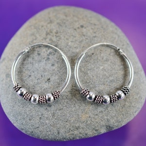 Peut inclure: Une paire de boucles d'oreilles créoles en argent avec de petites perles argentées. Les boucles d'oreilles sont sur un fond de pierre grise.