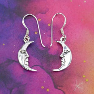 May include: A pair of silver-colored crescent moon earrings. Each earring features a detailed face carved into the moon shape. The earrings hang from a simple hook. The background is a colorful blend of purple, pink, and yellow.