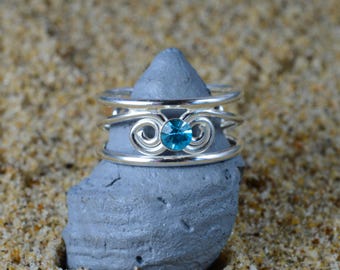 May include: A silver ring with a blue gemstone set in a swirl design. The ring is on a gray rock.