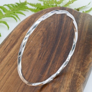 Könnte beinhalten: Ein silbernes Armreif-Armband mit einem gedrehten Design. Das Armband besteht aus glänzendem Metall und wird auf einer Holzoberfläche präsentiert. Der Armreif ist ein schlichtes, aber elegantes Schmuckstück.