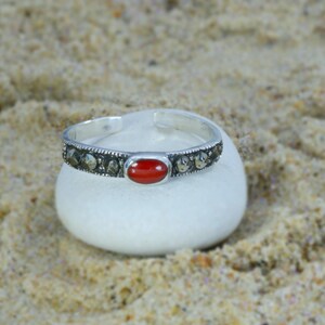 May include: A silver-toned cuff bracelet with a central oval red stone and surrounding small, dark stones. The bracelet rests on a white, rounded stone against a sandy background. Jewelry.