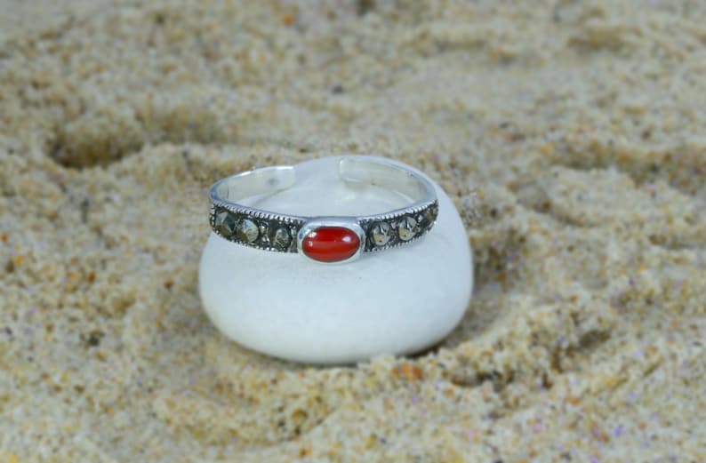 May include: A silver-toned cuff bracelet with a central oval red stone and surrounding small, dark stones. The bracelet rests on a white, rounded stone against a sandy background. Jewelry.