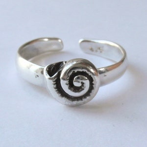 Puede incluir: Un anillo ajustable plateado con un diseño de concha en espiral. El anillo está abierto en la parte posterior y presenta una pieza central de concha texturizada y detallada. El anillo es probablemente de plata de ley.