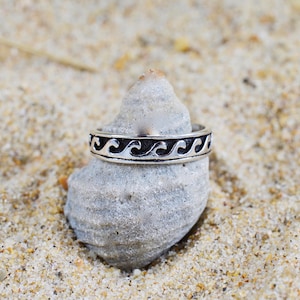 Può includere: Un anello d'argento con un motivo a onde, appoggiato su una pietra grigia su uno sfondo sabbioso. Il design dell'anello presenta una serie ripetuta di onde stilizzate in un colore scuro, creando un tema nautico.
