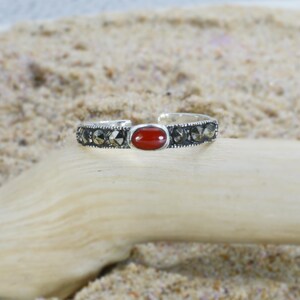May include: A silver-toned ring with a central oval, red gemstone. The band is detailed with small, dark stones. The ring is displayed on a piece of light-colored wood against a sandy background, showcasing a natural aesthetic.