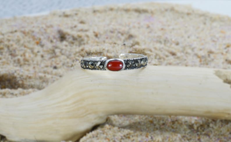 May include: A silver-toned ring with a central oval, red gemstone. The band is detailed with small, dark stones. The ring is displayed on a piece of light-colored wood against a sandy background, showcasing a natural aesthetic.