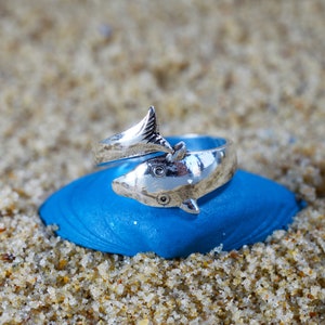 Puede incluir: Un anillo de plata con forma de delfín, con la cola envuelta alrededor de la banda. El anillo está apoyado sobre una superficie azul.