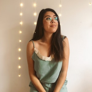 May include: A light green camisole and shorts set with a cream-colored lace trim. The camisole has thin straps and a scoop neckline. The shorts are visible at the bottom of the image. The person is wearing round, gold-rimmed glasses.