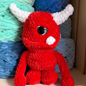 May include: A handmade red plush monster with white horns and a white spot. The monster has large black eyes and is sitting on a wooden shelf. The background features spools of yarn in shades of blue and green.