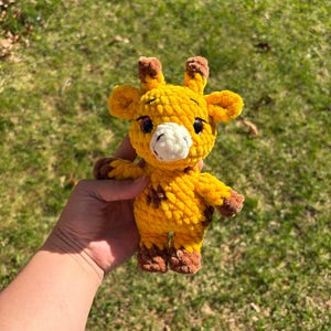 May include: A handmade, yellow crochet giraffe with brown spots, hooves, and antlers. The giraffe has a white muzzle and black eyes. The plush toy is held against a background of green grass.