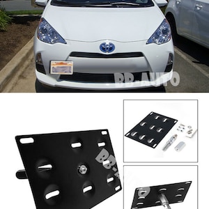 May include: A black metal license plate bracket with eight holes for mounting screws. The bracket is designed to fit a standard US license plate. The bracket is shown in front of a white Toyota Prius C.