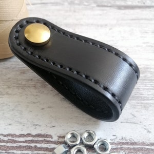 Pode incluir: Puxador de gaveta de couro preto com botão de metal dourado. O puxador tem uma borda costurada e é mostrado com um monte de porcas prateadas.