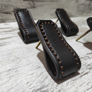 May include: Four brown leather drawer pulls with a decorative brown stitching detail. The pulls are attached to a white wooden surface with screws.