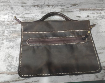 May include: A dark brown leather pouch featuring a top handle, a horizontal zippered pocket on the front, and visible cream-colored stitching along the edges. The bag has a rectangular shape with rounded corners and a dark brown zipper pull.
