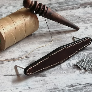 Könnte beinhalten: Ein brauner Ledergriff mit Nähten und zwei Metallschrauben. Eine Spule mit beigefarbenem Garn, ein Holz-Stechbeitel und ein Haufen silberner Muttern sind ebenfalls sichtbar.