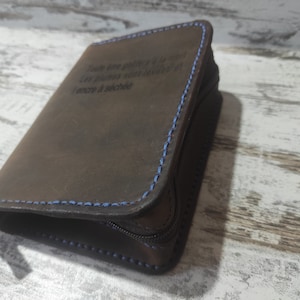 Puede incluir: Una cartera de cuero marr&oacute;n oscuro con costuras azules en contraste y cierre de cremallera. El frente est&aacute; grabado con el texto en franc&eacute;s: Toute &acirc;me go&ucirc;tera &agrave; la mort. Les plumes sont lev&eacute;es et l'encre a s&eacute;ch&eacute;e. Se observa un peque&ntilde;o detalle met&aacute;lico color lat&oacute;n.