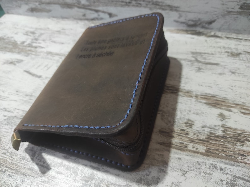 Puede incluir: Una cartera de cuero marr&oacute;n oscuro con costuras azules en contraste y cierre de cremallera. El frente est&aacute; grabado con el texto en franc&eacute;s: Toute &acirc;me go&ucirc;tera &agrave; la mort. Les plumes sont lev&eacute;es et l'encre a s&eacute;ch&eacute;e. Se observa un peque&ntilde;o detalle met&aacute;lico color lat&oacute;n.