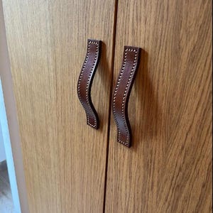 Peut inclure: Deux poignées de porte de placard en cuir marron avec des détails de couture. Les poignées sont fixées à une porte de placard en bois.