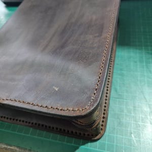 Puede incluir: Un estuche de cuero marr&oacute;n oscuro con un acabado mate suave y costuras visibles a lo largo de los bordes. El art&iacute;culo rectangular cuenta con un cierre de cremallera en el lateral, mostrando un dise&ntilde;o estructurado con costuras limpias.