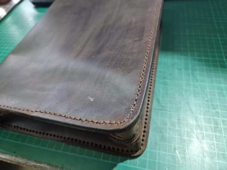 Puede incluir: Un estuche de cuero marr&oacute;n oscuro con un acabado mate suave y costuras visibles a lo largo de los bordes. El art&iacute;culo rectangular cuenta con un cierre de cremallera en el lateral, mostrando un dise&ntilde;o estructurado con costuras limpias.