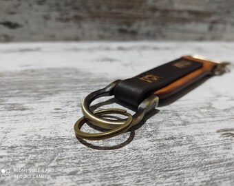 May include: A handcrafted leather keychain with a dark brown top layer and a lighter brown bottom layer. The keychain features a brass-colored clasp and key rings. The leather is embossed with a design. The keychain is designed for holding keys.