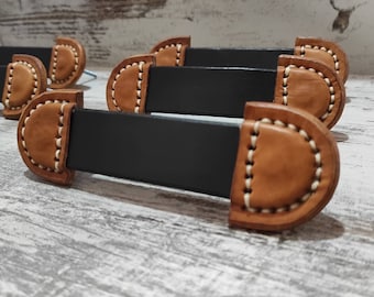 Puede incluir: Cuatro tiradores de cajón de cuero marrón con inserciones de metal negro y costuras blancas. Los tiradores están dispuestos en una fila sobre una superficie de madera clara.