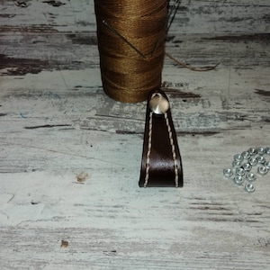 May include: A brown leather handle with a white stitch detail and a silver metal button. The handle is on a white wooden surface with a spool of thread and metal nuts.