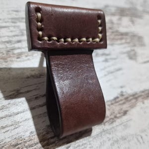 Könnte beinhalten: Ein brauner Leder-Schlaufen-Griff mit weißen Nähten. Der Griff ist so konzipiert, dass er an einer Tasche oder einem anderen Gegenstand befestigt werden kann.