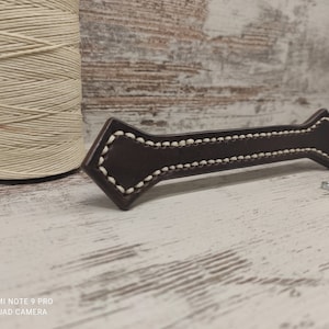 May include: A dark brown leather handle with white stitching. The handle has a diamond-shaped design on each end and is likely for a drawer or cabinet. A spool of beige thread and several nuts are also visible.
