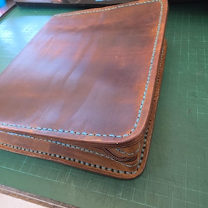 Puede incluir: Una carpeta de cuero marr&oacute;n con cremallera y costuras turquesa visibles a lo largo de los bordes. El cuero tiene un acabado liso y mate con variaciones de color naturales, y una cremallera marr&oacute;n recorre el lateral de la cubierta.