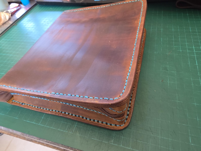 Puede incluir: Una carpeta de cuero marr&oacute;n con cremallera y costuras turquesa visibles a lo largo de los bordes. El cuero tiene un acabado liso y mate con variaciones de color naturales, y una cremallera marr&oacute;n recorre el lateral de la cubierta.