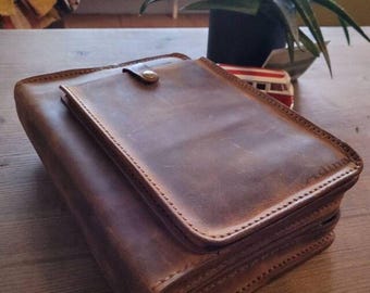 May include: A rectangular brown leather case with a zippered closure and a smaller flap pocket on the top secured by a single metal snap button. The leather has a smooth, matte finish with visible stitching along the edges and a small embossed logo.