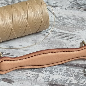 Könnte beinhalten: Ein hellbrauner Ledergriff mit einer genähten Kante und zwei Schrauben. Der Griff befindet sich auf einer weißen Holzoberfläche mit einer Spule beigefarbenen Garns und einer Nadel.