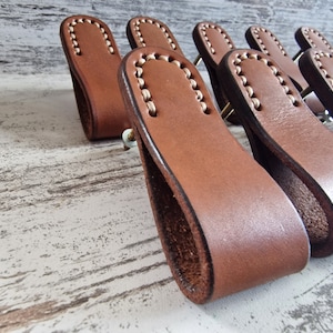 Op de afbeelding: Een rij van bruine lederen ladegrepen met witte stiksels. De grepen zijn in een verspringend patroon gerangschikt en hebben een licht gebogen vorm.