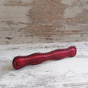 Könnte beinhalten: Ein roter Leder-Schrankgriff mit wellenförmigem Design und dekorativen Nähten.