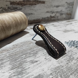 May include: A brown leather pull with a white stitch detail and a gold metal knob. The pull is on a white wooden surface.