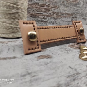 Può includere: Una maniglia in pelle marrone chiaro con due rivetti in metallo dorato. La maniglia è cucita con un filo marrone chiaro. Ci sono diversi rivetti in metallo dorato e argentato su una superficie di legno.