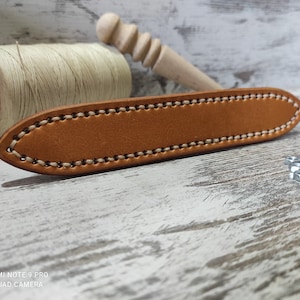 Può includere: Primo piano di un cinturino in pelle marrone con cuciture bianche. Il cinturino è appoggiato su una superficie di legno.