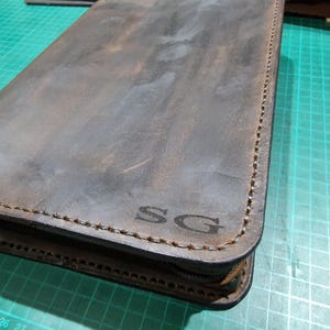 Puede incluir: Un estuche rectangular de cuero marr&oacute;n oscuro con esquinas redondeadas y costuras visibles a lo largo de los bordes. La superficie presenta una textura desgastada y las letras SG est&aacute;n grabadas en la esquina inferior derecha.