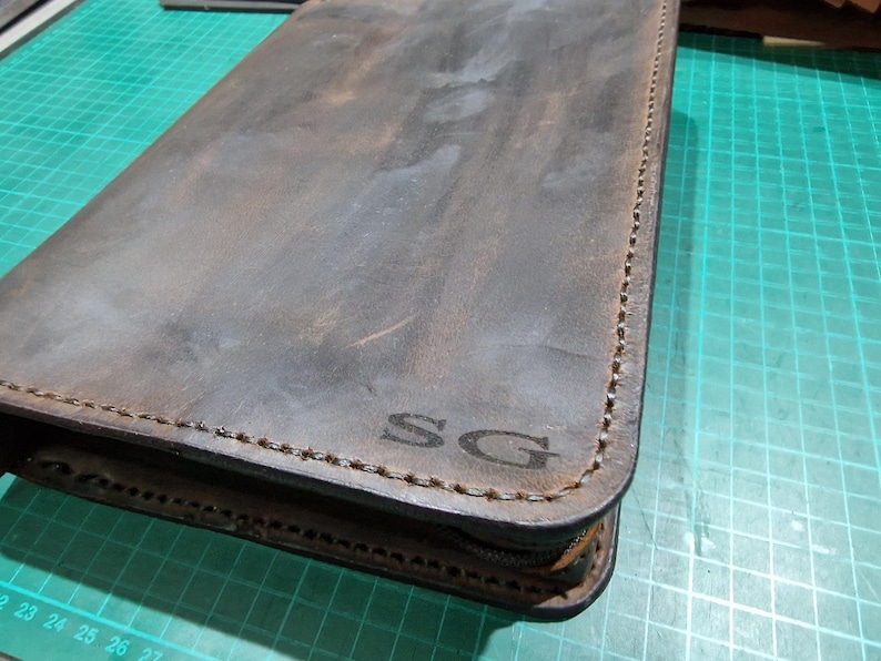 Puede incluir: Un estuche rectangular de cuero marr&oacute;n oscuro con esquinas redondeadas y costuras visibles a lo largo de los bordes. La superficie presenta una textura desgastada y las letras SG est&aacute;n grabadas en la esquina inferior derecha.