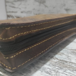 Puede incluir: Un estuche rectangular de cuero marr&oacute;n con una cremallera de color marr&oacute;n oscuro a lo largo del lateral. Los bordes del cuero est&aacute;n unidos con costuras visibles de color amarillo dorado que recorren el per&iacute;metro.