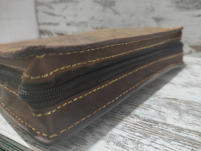 Puede incluir: Un estuche rectangular de cuero marr&oacute;n con una cremallera de color marr&oacute;n oscuro a lo largo del lateral. Los bordes del cuero est&aacute;n unidos con costuras visibles de color amarillo dorado que recorren el per&iacute;metro.