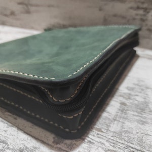 Puede incluir: Un estuche de cuero verde oscuro con acabado mate y costuras de color claro visibles en los bordes. El objeto rectangular cuenta con una cremallera negra a lo largo del lateral, asegurando el contenido dentro del marco de cuero estructurado.