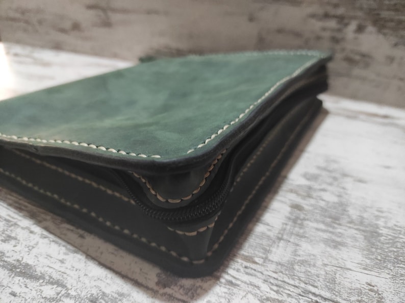 Puede incluir: Un estuche de cuero verde oscuro con acabado mate y costuras de color claro visibles en los bordes. El objeto rectangular cuenta con una cremallera negra a lo largo del lateral, asegurando el contenido dentro del marco de cuero estructurado.