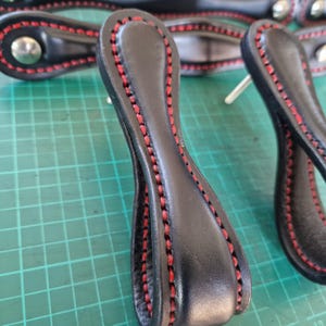 Può includere: Tre tiretti per cassetti in pelle nera con cuciture rosse. I tiretti hanno un design a forma di anello e sono fissati a una base in metallo.