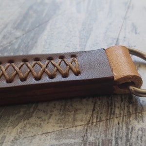 Peut inclure: Un porte-clés en cuir marron avec une boucle en cuir beige et un anneau en métal argenté. Le cuir est cousu avec du fil marron en forme de croix.