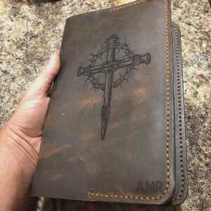Puede incluir: Una funda de cuero marr&oacute;n oscuro para diario con un dise&ntilde;o grabado a l&aacute;ser de una cruz envuelta en una corona de espinas. La funda tiene costuras visibles en color canela a lo largo de los bordes y las iniciales AMR en la esquina inferior.