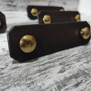 Può includere: Primo piano di una maniglia per cassetto in pelle nera con due pomelli in metallo dorato. La maniglia è su una superficie in legno sbiancato.