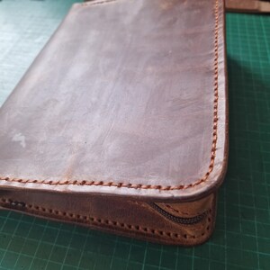 Puede incluir: Un estuche rectangular de cuero marr&oacute;n con bordes cosidos a mano con hilo grueso. El estuche presenta una textura de superficie lisa y mate, junto con una cremallera de color marr&oacute;n oscuro en el lateral para cerrar el contenido de forma segura.
